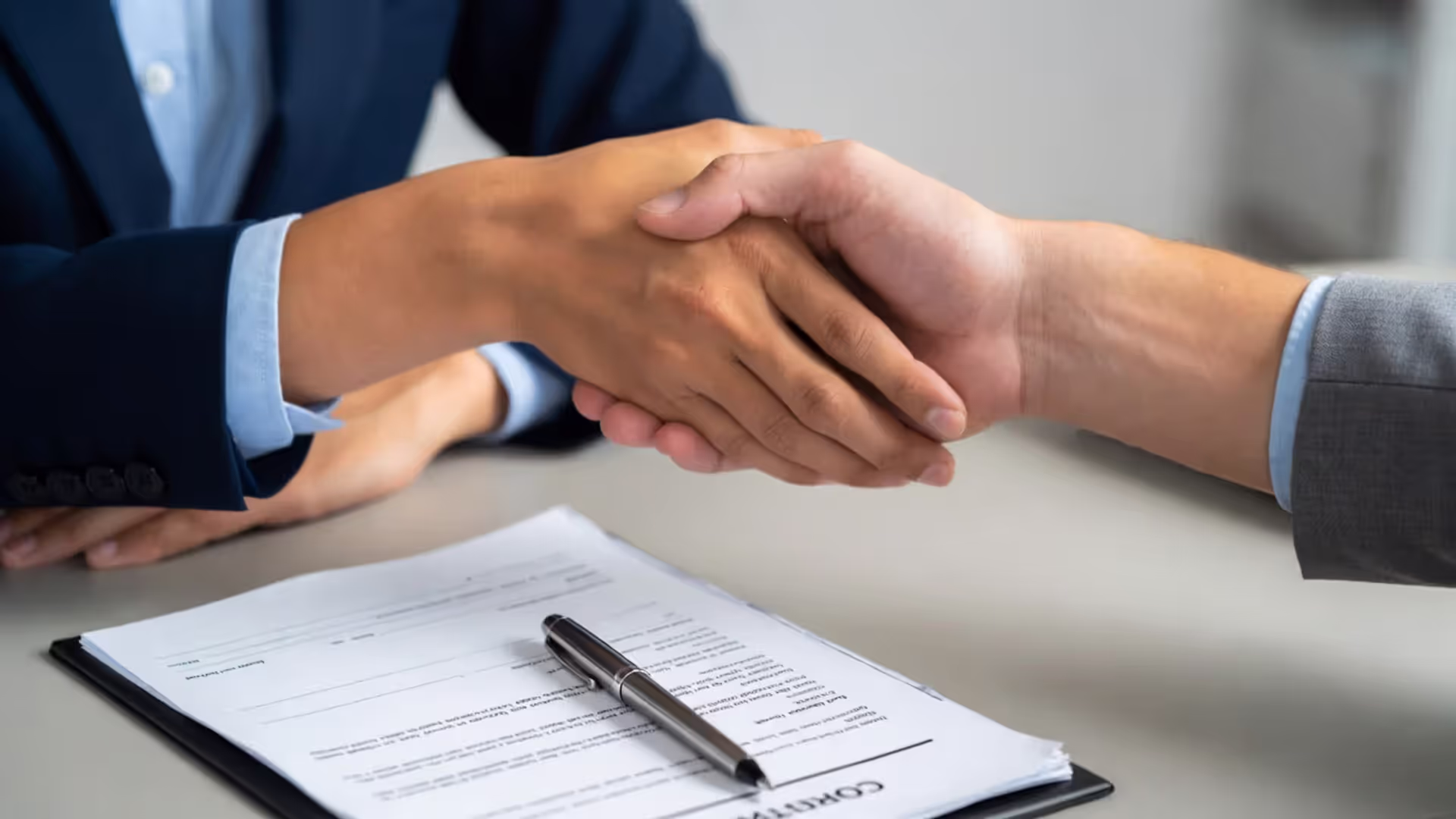 Two people shaking hands over a contract document with a pen on the page.
